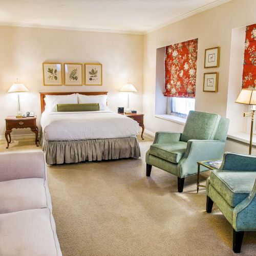 A cozy hotel suite with a king bed, two nightstands, a sofa, two teal chairs, side lamps, framed art, and red floral curtains by the windows.