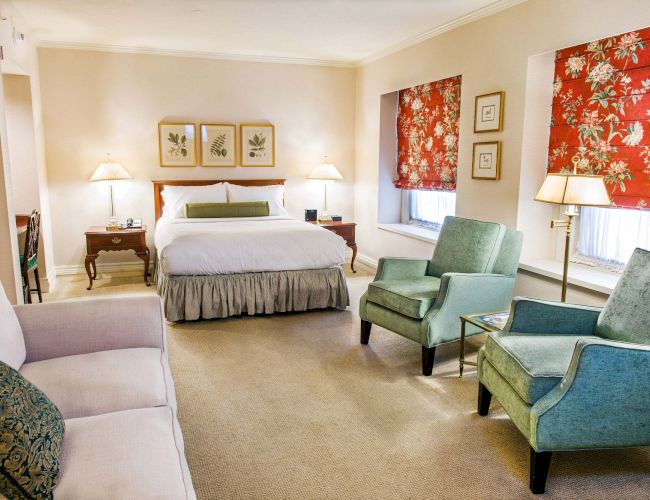 A cozy hotel suite with a king bed, two nightstands, a sofa, two teal chairs, side lamps, framed art, and red floral curtains by the windows.