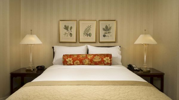 A neatly made hotel bed with white linens, beige bedspread, two lamps, and botanical wall art above the headboard, framed pictures.