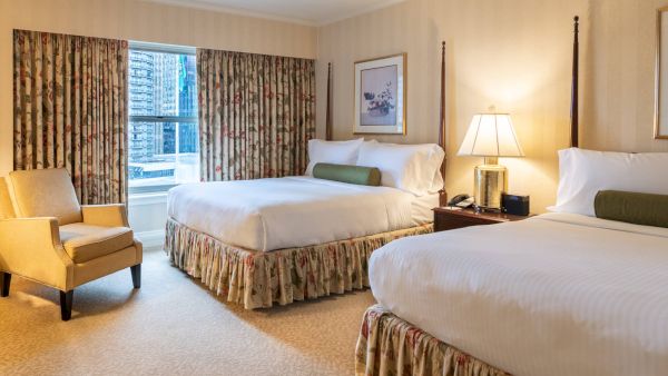 A hotel room with two queen beds, floral bed skirts, beige walls, a nightstand with a lamp, a chair by the window, and draped curtains.