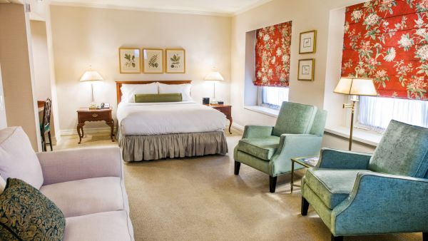 A deluxe hotel suite with a king bed, two green armchairs, a sofa, lamps, and floral red window coverings, creating a cozy, classic room.