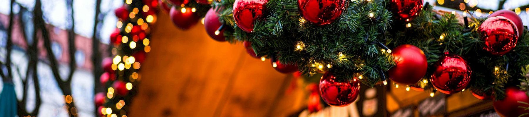A festive market stall decorated with evergreen garlands and red baubles, twinkling lights framing the wooden booth at dusk.