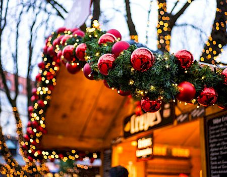 A festive market stall decorated with evergreen garlands and red baubles, twinkling lights framing the wooden booth at dusk.