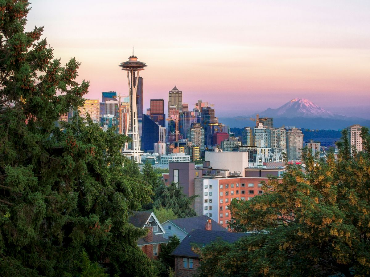 A city skyline with the Space Needle, mid-rise buildings, and trees framing the scene at sunset, creating a warm, scenic urban view.
