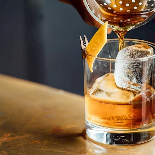A glass of whiskey on the rocks with a large ice cube and an orange peel garnish being poured into it.