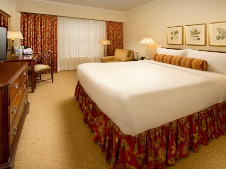 Classic King hotel room featuring decorative pillows, framed botanical art, a seating area, desk and TV, warm lighting, and patterned drapes.