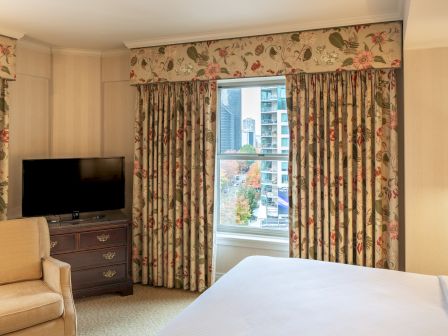 A cozy hotel room with floral curtains, a flat-screen TV on a dresser, a light armchair, and a window showing a cityscape outside, ending in a period.