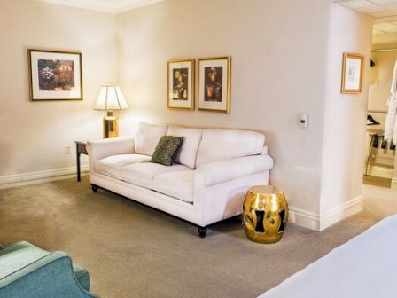 A cozy living room with a white sofa, green armchairs, framed art, a side table lamp, and a gold decorative stool near a doorway.