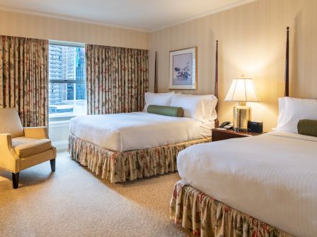Two double beds with floral bedskirts, a nightstand lamp, a chair by the window, and a window view in a cozy hotel room.