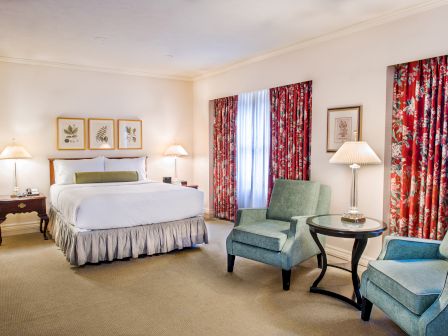A hotel suite with a king bed, two blue armchairs, side tables, lamps, and red floral curtains in a bright, neutral room. Only a cozy, tidy space.