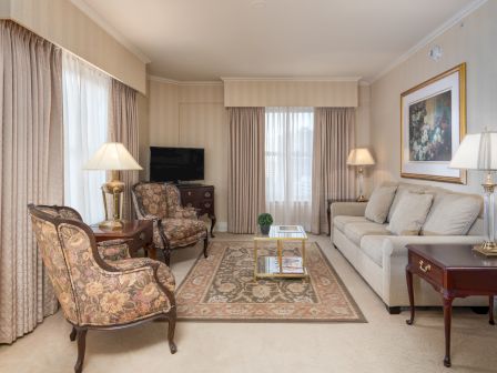 A elegant living room with beige walls, light drapes, a plush sofa, patterned armchairs, a coffee table on a decorative rug, and lamps on side tables.