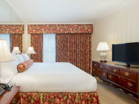 A hotel room with a large bed, matching floral curtains and bed skirt, wooden dresser with a TV, nightstands, and lamps on both sides, warm lighting.