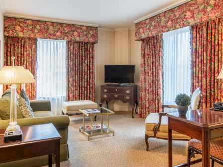 A classic hotel suite: floral drapes, two windows, green sofa, wooden desk, lamps, a small TV on a cabinet, and a cozy seating area with a coffee table.