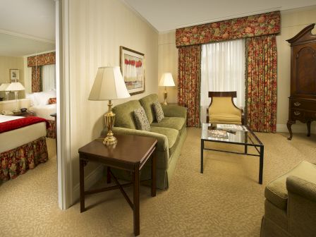 A hotel suite with a sitting area, sofa, coffee table, lamps, armoire, and two beds visible in the adjoining room, cozy and traditional.