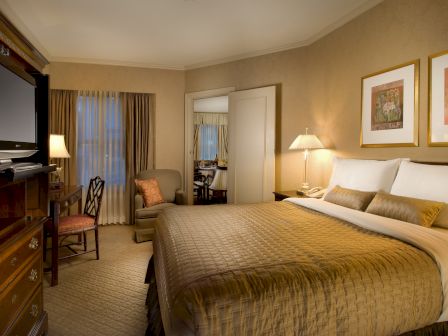 A hotel room with a large bed, lamps, framed art, a desk and chair, a dresser with TV, and a seating area near a window.