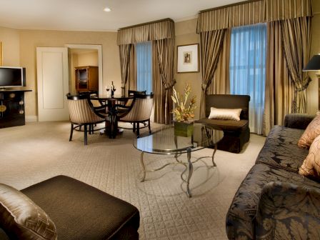 A luxurious hotel suite with a seating area, round glass coffee table, patterned sofa, armchairs, curtains, and a dining table in the background, warm lighting.