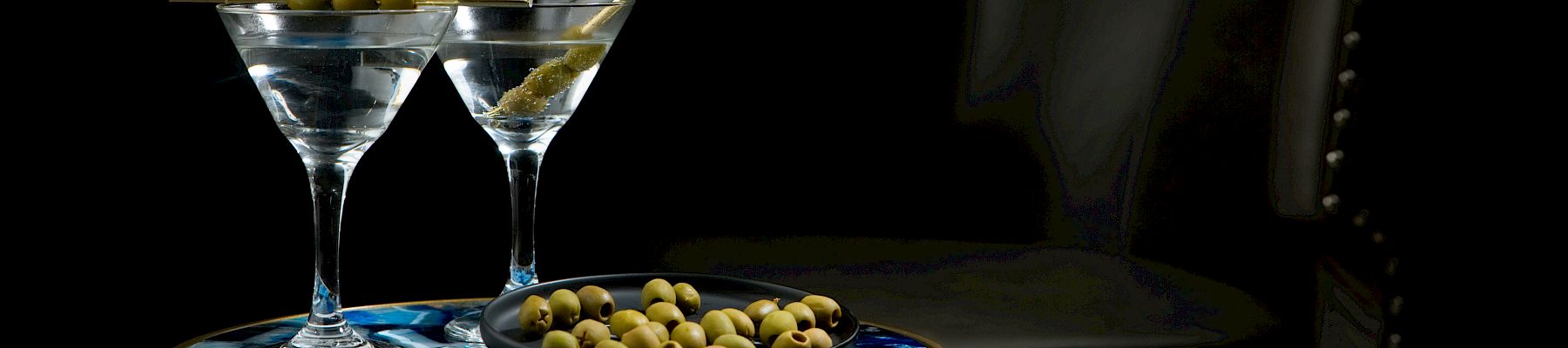 Two martini cocktails in glasses with green olives sit on a blue tray beside a black bottle in dim light.