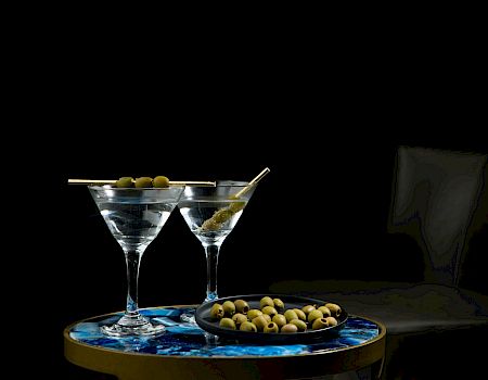 Two martini cocktails in glasses with green olives sit on a blue tray beside a black bottle in dim light.