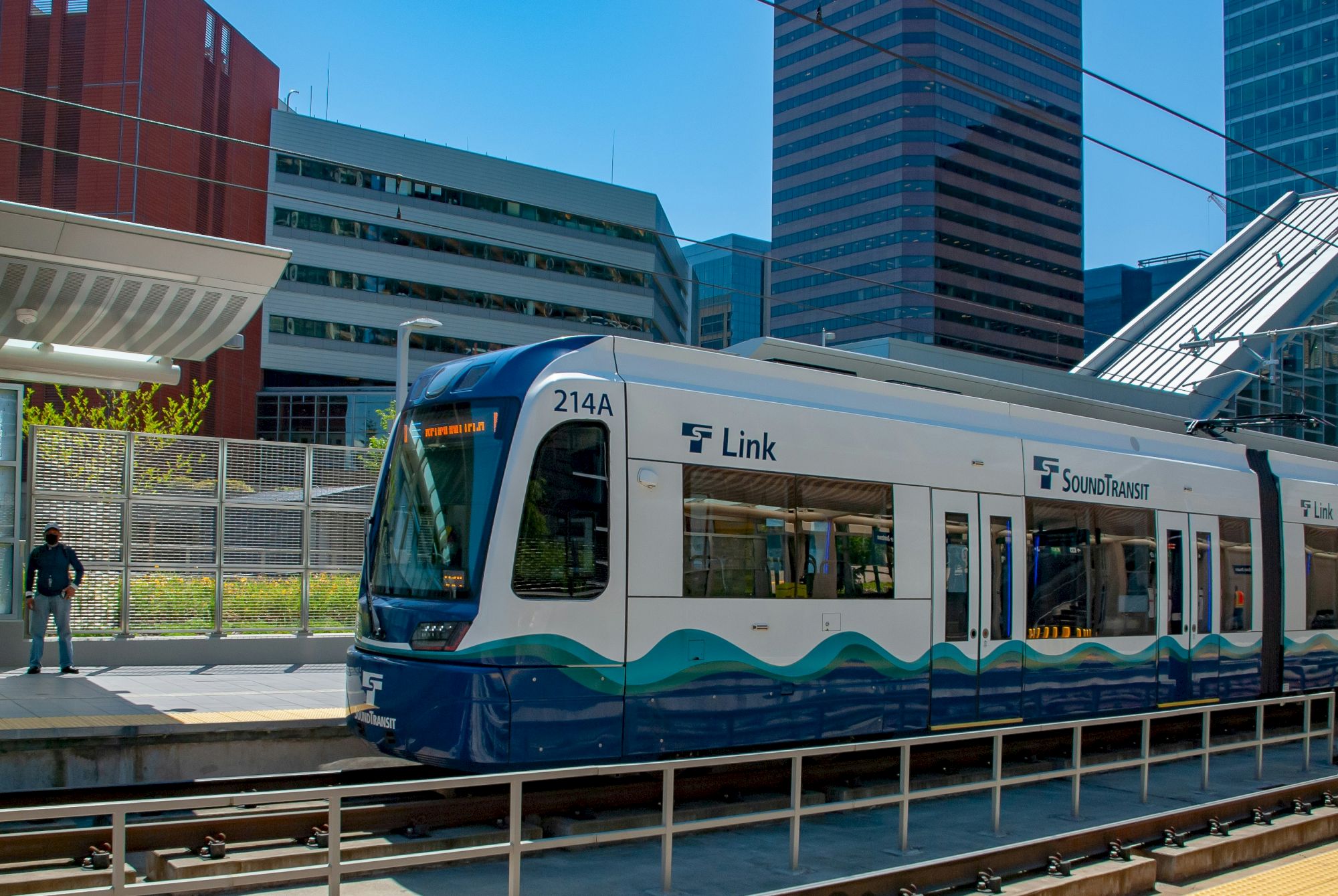 A light rail train at a sunlit station with blue-green wave designs, modern buildings in the background, and a pedestrian nearby.