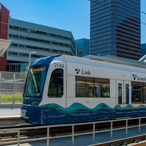 A modern light rail train at a station with blue wave graphics, urban glass buildings in the background, and a pedestrian on the platform.