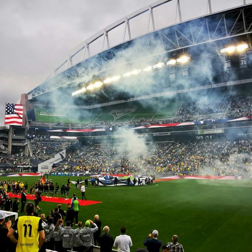 A crowded football stadium on game day with smoke/flares, a large screen, and fans waving flags in a green field arena.