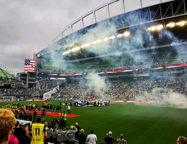 A crowded football stadium on game day with smoke/flares, a large screen, and fans waving flags in a green field arena.