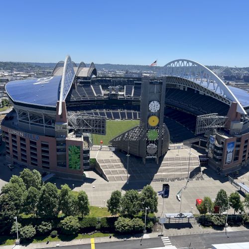 Aerial view of a large modern stadium with a curved roof, trees lining the front, and a clear blue sky above, lots of open seating and surrounding roads.