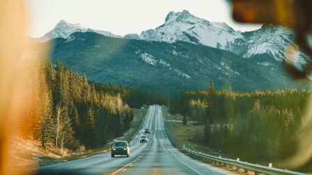 A long highway through a forested valley with mountains in the background and a few cars driving toward the distance.