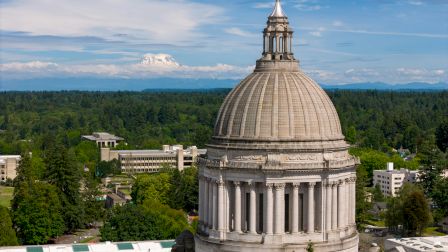 The image shows a large domed capitol-style building with a tall dome and classical columns, set among trees and smaller structures.