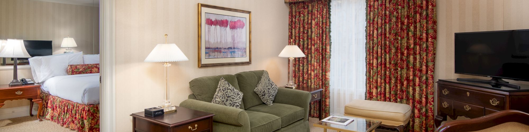 A hotel living area with a green sofa, twin lamps, framed wall art, a side table, floral curtains, a TV on a wooden cabinet, and a doorway to a bedroom.