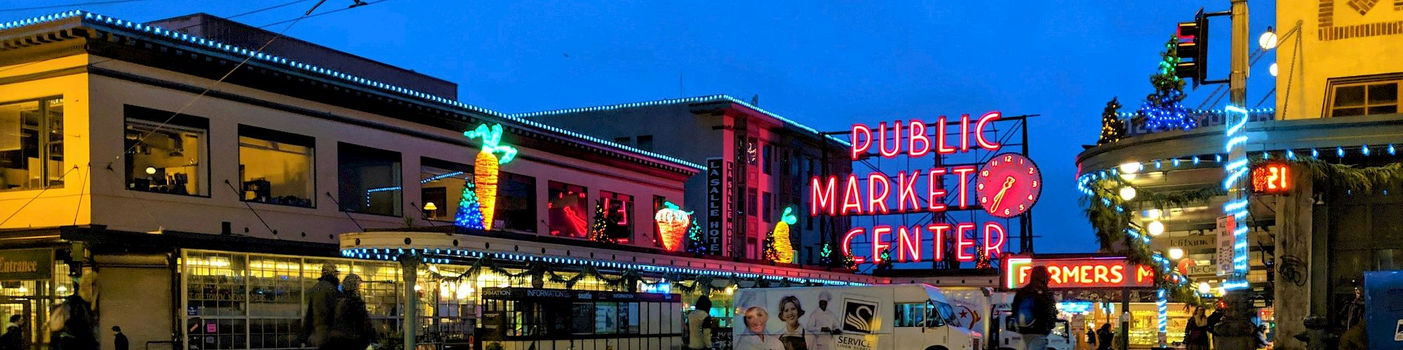 Neon-lit Public Market Center at dusk, with holiday lights and a parade bus on a plaza.