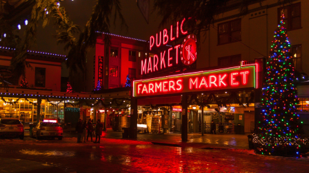 A festive night scene outside &ldquo;Public Market&rdquo; with neon &ldquo;Farmers Market&rdquo; lights, twinkling string decor, and a Christmas tree.