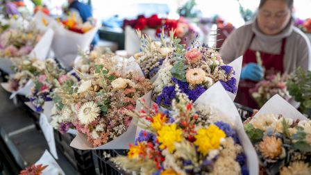A flower shop table crowded with colorful bouquets as a customer selects blooms in the background.