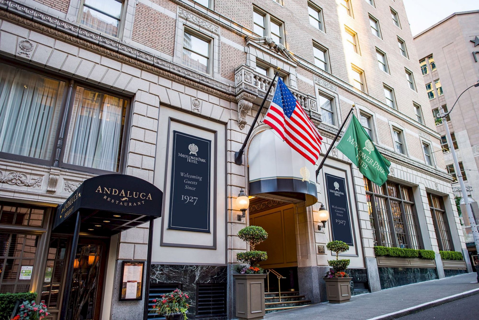A tall, elegant hotel with an American flag and a green flag outside, a welcoming entrance, and stylish fa&ccedil;ade on a city street.