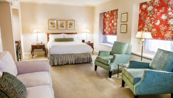 A hotel suite with a king bed, a sofa, two teal chairs, side tables, lamps, floral red curtains, and framed art on beige walls.