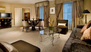 A hotel suite living area with a sofa, armchairs, glass coffee table, dining table, window drapes, flat-screen TV, and warm beige d&eacute;cor, cozy and elegant.