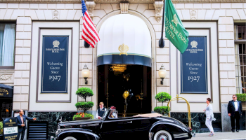 A black vintage car parks in front of a grand hotel entrance with blue banners, flags, and well-dressed people under an arched doorway.