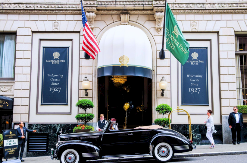 A black vintage car parks in front of a grand hotel entrance with blue banners, flags, and well-dressed people under an arched doorway.
