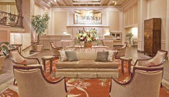 A luxurious hotel lobby with a beige sofa set, several cushioned chairs, a central floral arrangement, warm lighting, plants, and a grand reception area in the background.