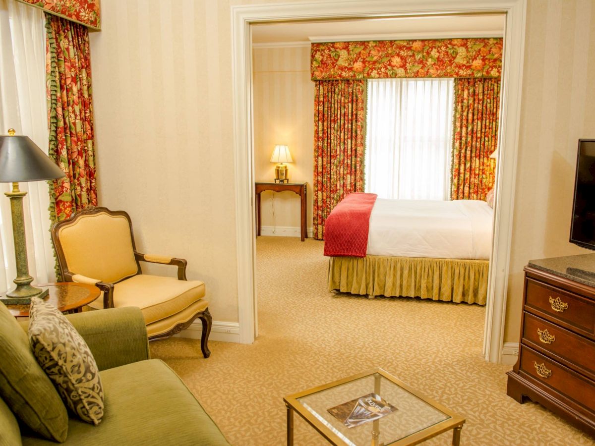 A hotel suite with a living area, green sofa, yellow chair, glass coffee table, and a separate bedroom with floral drapes and a bed with red throw, plus a dresser and TV.