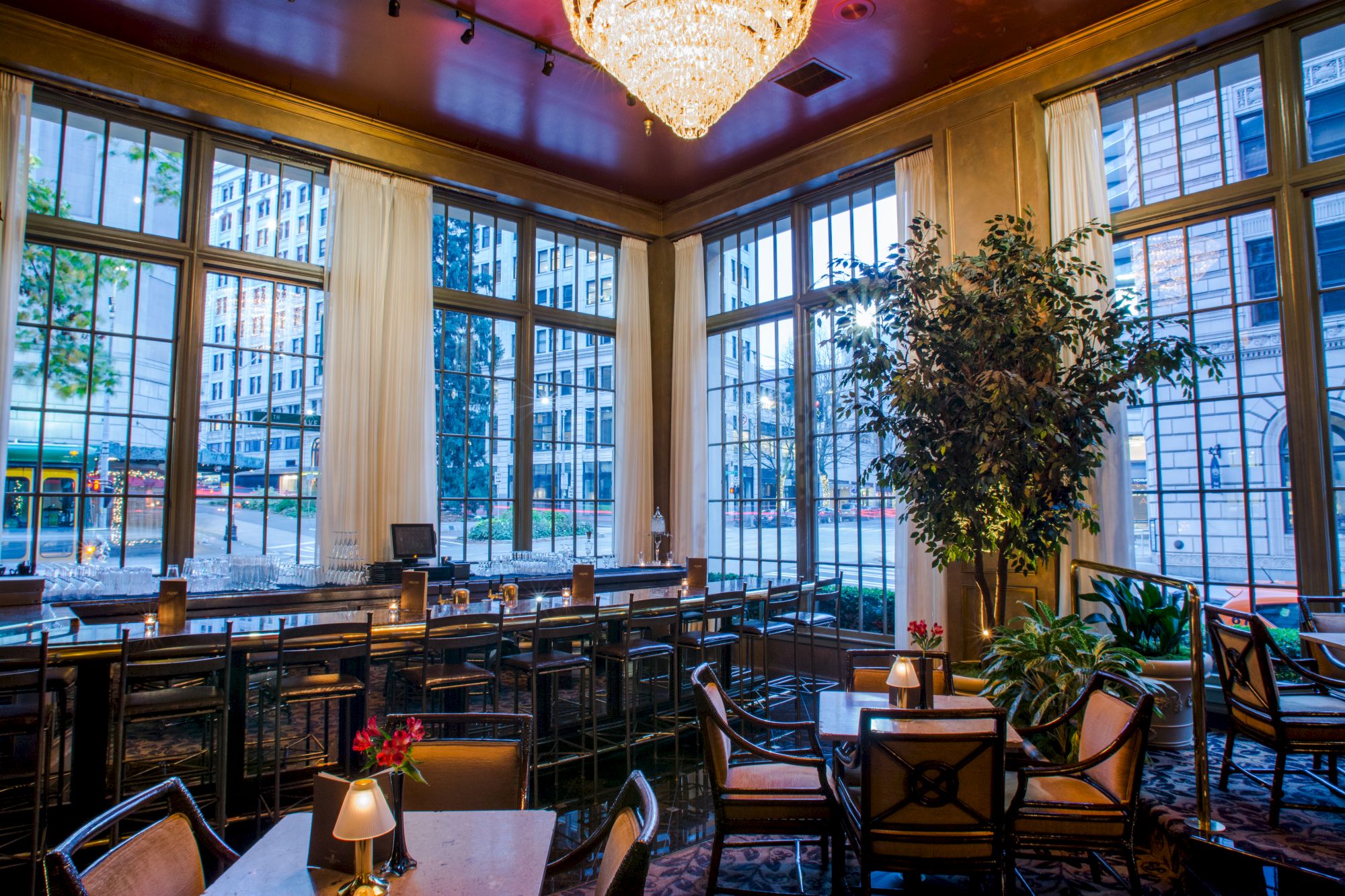 A stylish indoor cafe or restaurant with large windows, tables, chairs, a chandelier, and a potted plant in warm lighting, with a city view.