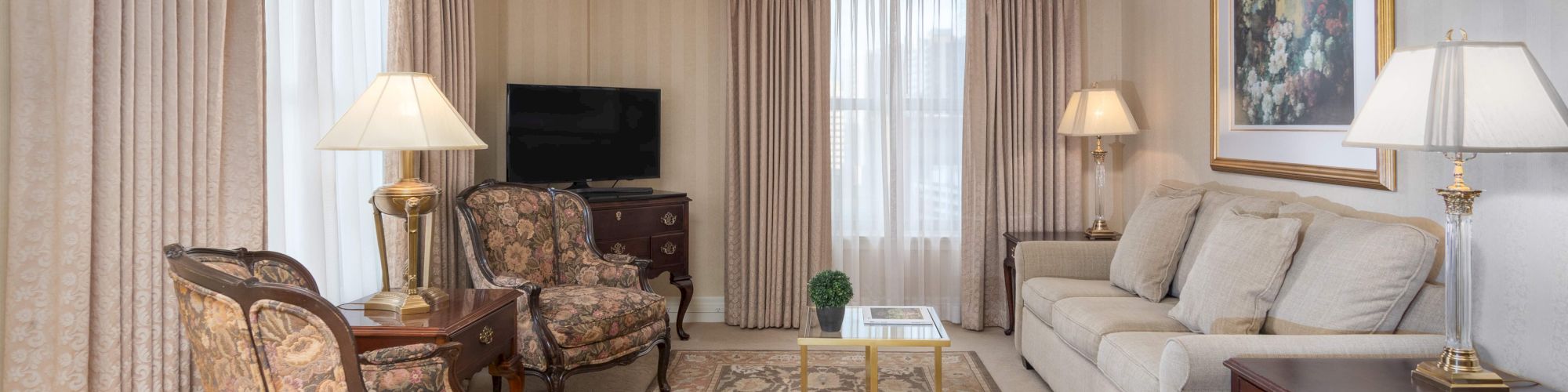 A luxurious living room with beige walls, cream sofa, patterned armchairs, wooden coffee table, area rug, side tables with lamps, and a framed artwork above the sofa.