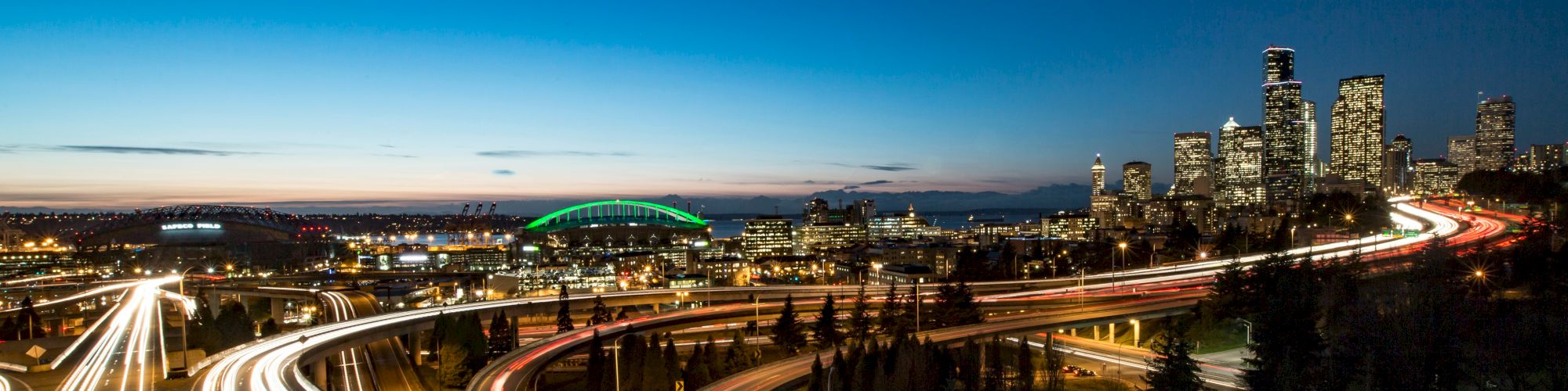 A nighttime cityscape with a glowing skyline, winding highways with streaking car lights, and a distant green bridge under a dark blue sky.