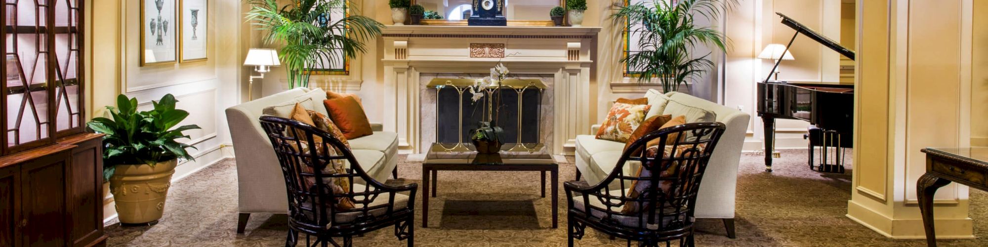 A cozy hotel lobby with a central fireplace, comfortable armchairs, a piano on the right, potted plants, framed art, and warm ambient lighting.