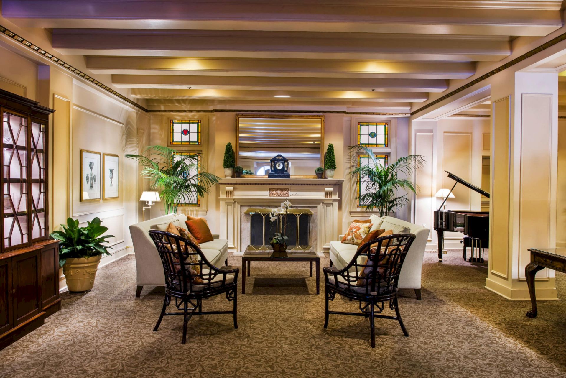 A cozy hotel lobby with a central fireplace, cream sofas, wicker chairs, potted plants, warm lighting, and a grand piano to the right.