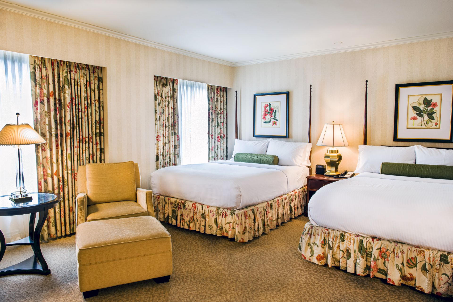 A bright hotel room with two neatly made beds, floral curtains, framed botanical artwork, and warm bedside lamps for a cozy stay.