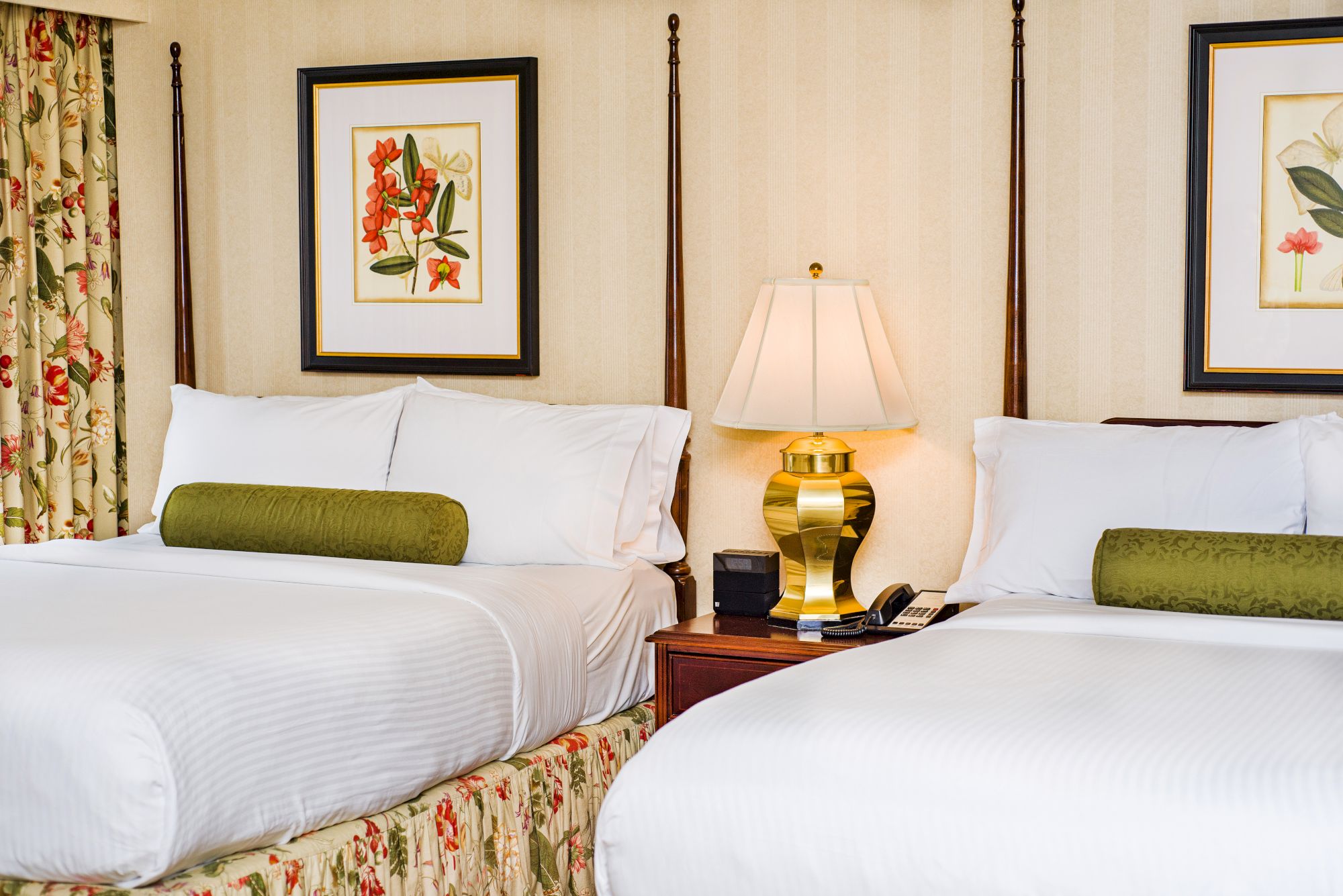 Two neatly made beds in a hotel room with a nightstand, lamp, framed botanical art, and floral-patterned curtains; cozy and bright.