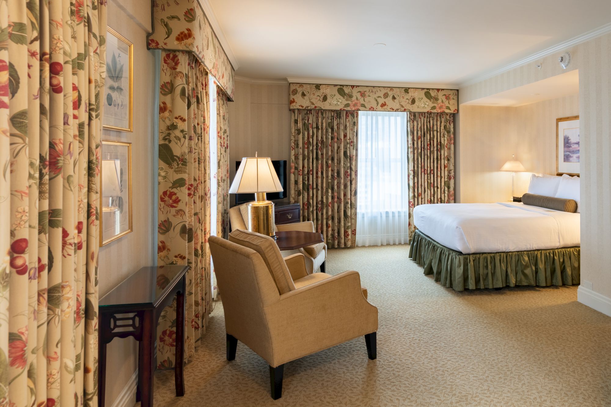 A hotel suite with floral curtains, a desk, two chairs, a lamp, and a king bed; warm lighting, neutral tones, and a window with drapes.