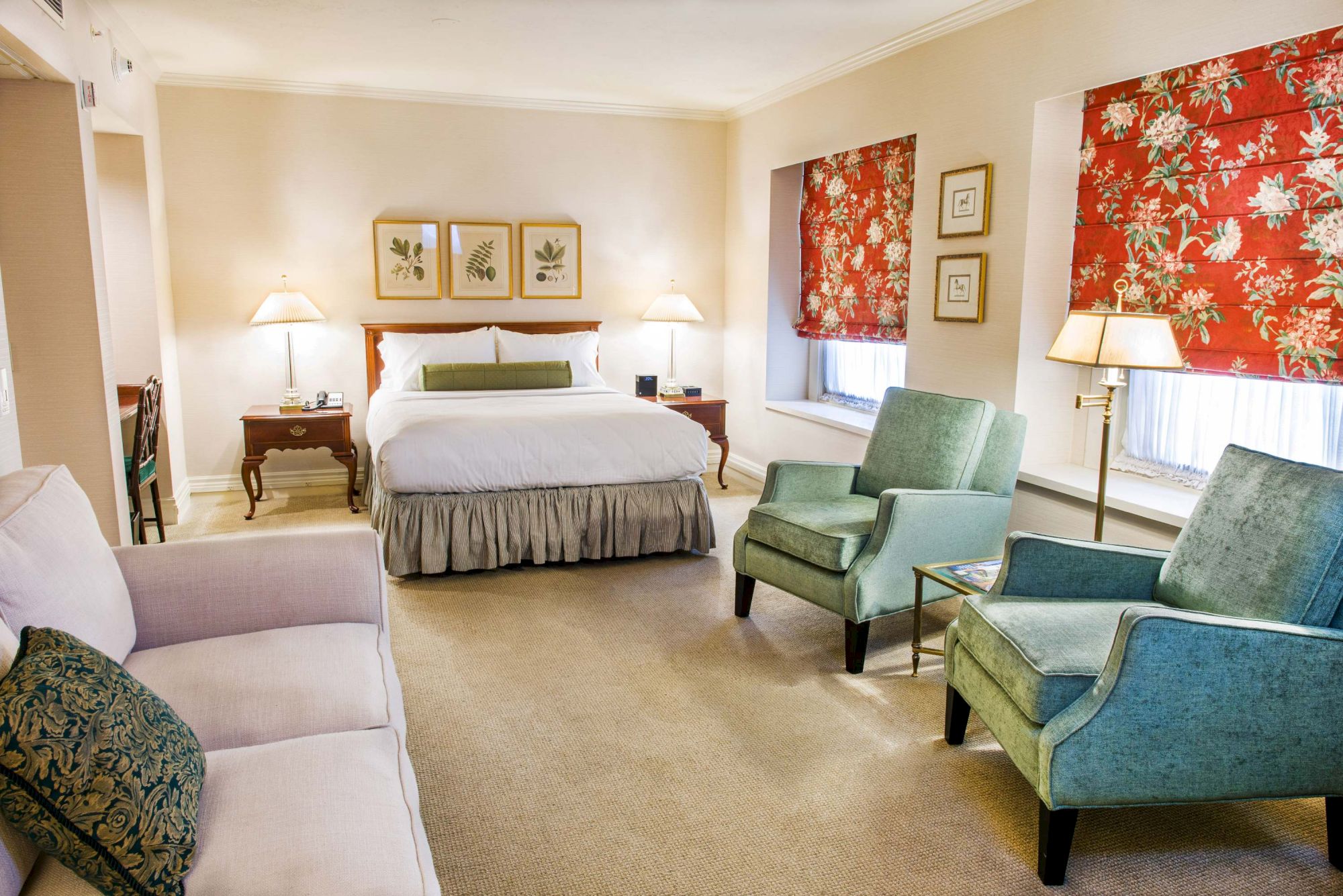 A bright hotel suite with a king bed, two nightstands, a sofa, two teal armchairs, and red floral curtains, plus lamps and framed art.
