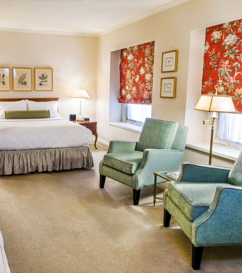 A hotel room with a bed, two green chairs, and a window with floral curtains.